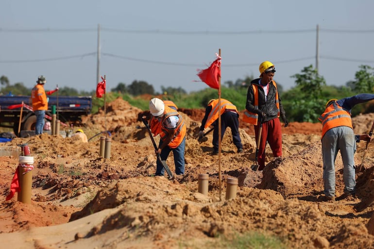 Seis trabajadores en un sitio de construcción, con cascos amarillos y chalecos naranjas, excavando en terreno de tierra.
