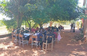 Grupo de jóvenes sentados al aire libre bajo un árbol, algunos escribiendo, otros discutiendo, con ambiente informal de estudio.