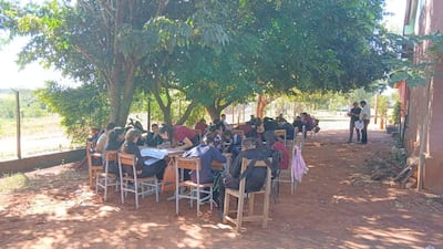Grupo de jóvenes sentados al aire libre bajo un árbol, algunos escribiendo, otros discutiendo, con ambiente informal de estudio.