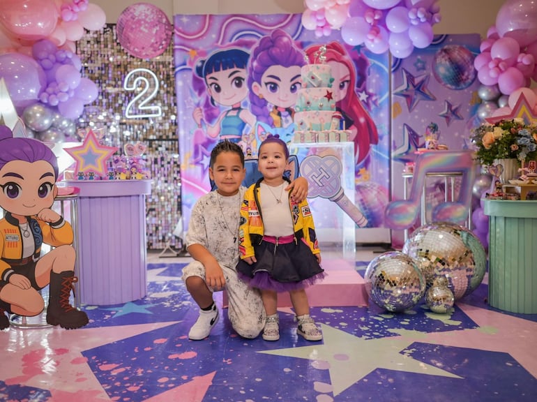Niño con conjunto claro y niña con abrigo colorido, sonrientes en fiesta de cumpleaños con globos lilas y pastel decorado.