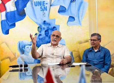 Hombre canoso en camisa clara habla, mientras otro hombre con gafas y camisa azul lo escucha atentamente en un ambiente formal.