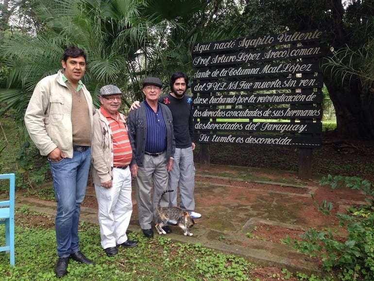 Cinco hombres en un parque con vegetación, conversando y reflexionando, mientras un gato se encuentra entre ellos.