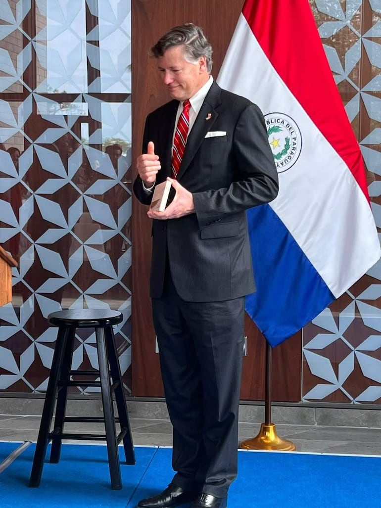 Christopher Landau de pie en traje negro y corbata de rayas, frente a la bandera de Paraguay en un ambiente formal.