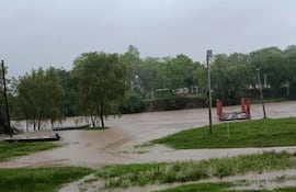 Paisaje inundado con césped y árboles verdes. Botes anclados y estructura metálica roja en el entorno ribereño.