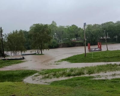 Paisaje inundado con césped y árboles verdes. Botes anclados y estructura metálica roja en el entorno ribereño.