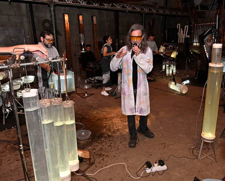 Hombre canoso con abrigo de laboratorio habla al micrófono, mientras otros manipulan instrumentos y agua en un ambiente artístico.