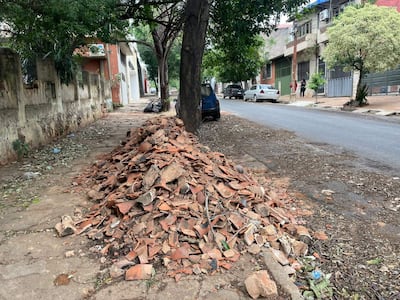 Montículo de tejas rotas en la vereda con árboles y vehículos en el fondo.