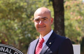 González Morel con blazer oscuro y corbata roja, sonriente, frente a un emblema institucional en un entorno verde.
