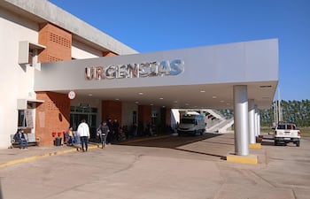 Entrada del hospital con letrero de 'URGENCIAS'. Personas esperando, un hombre de pie hablando por teléfono, cielo despejado.