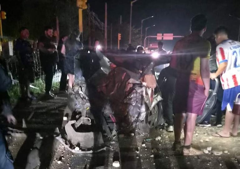 Multitud observa un vehículo gravemente dañado; camisetas de diferentes colores en la escena, y personas conversando en un entorno urbano.