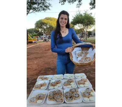 Diana Cañete, mujer de pie con camisa azul y jeans, sostiene canasta de pasteles frente a bandejas llenas en un parque.