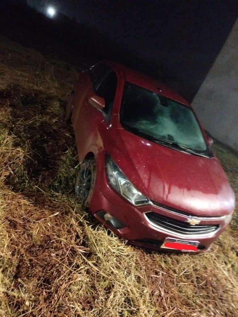 Automóvil Chevrolet rojo atascado en hierba alta y húmeda, con poca iluminación ambiental de fondo.