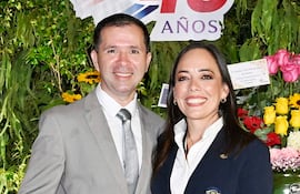 Hombre en traje claro y mujer en blazer oscuro sonríen en un ambiente festivo decorado con plantas y flores.