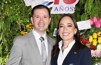 Hombre en traje claro y mujer en blazer oscuro sonríen en un ambiente festivo decorado con plantas y flores.