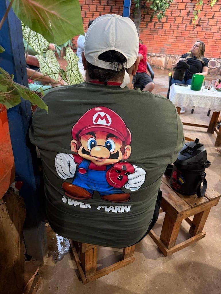 Hombre de espaldas con camiseta de Super Mario sentado en un banco, rodeado de personas en ambiente casual y verde.