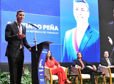 Santiago Peña en traje oscuro frente a un micrófono, mientras una mujer de rojo lo escucha atentamente.