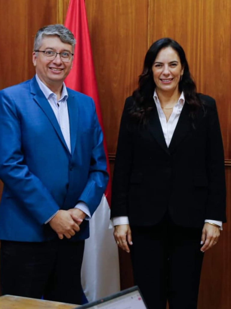 Hombre canoso en chaqueta azul y mujer de cabello oscuro en camisa blanca, sonríen frente a un fondo de banderas paraguayas.