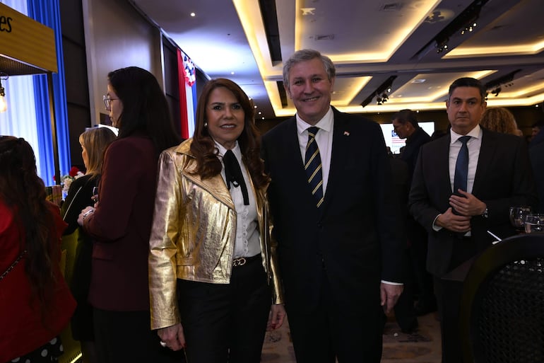 Mujer de cabello castaño claro con chaqueta dorada sonríe junto a un hombre canoso en traje oscuro en un evento interior.