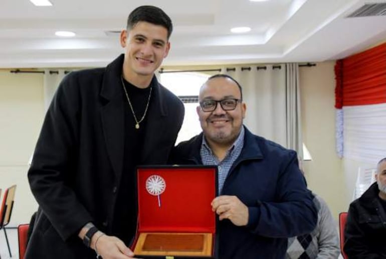 Hombre alto en camiseta negra recibe un reconocimiento de otro hombre con abrigo oscuro y camisa a cuadros, ambos sonrientes en un ambiente formal.