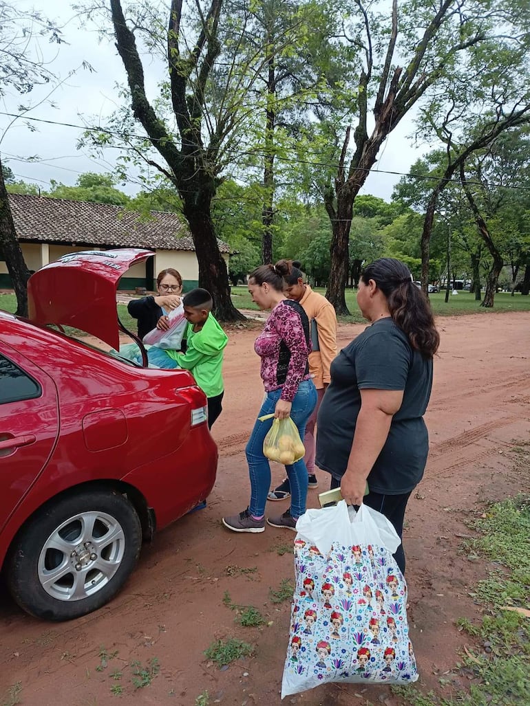 Mujer entrega objeto a un niño con camiseta verde, mientras otro niño observa y una mujer sostiene una bolsa, rodeados de árboles.