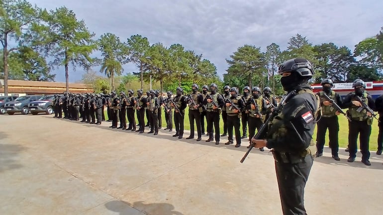 Agentes de seguridad uniformados en formación, con armas largas, en un ambiente exterior con árboles y camino pavimentado.