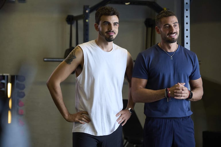 Hombres en un gimnasio, uno viste camiseta blanca y tatuaje, el otro camiseta azul, ambos expresan alegría y se comunican.