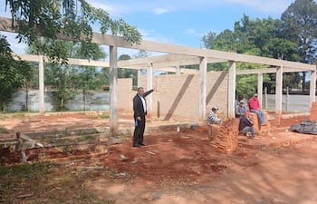 Hombre en traje oscuro señala hacia la construcción; otros cuatro conversan sobre ladrillos en un ambiente de obra exterior.