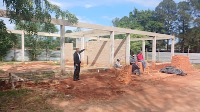 Hombre en traje oscuro señala hacia la construcción; otros cuatro conversan sobre ladrillos en un ambiente de obra exterior.