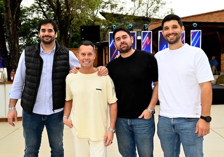Cuatro hombres sonrientes en camisetas amarillas y negras posan en un elegante ambiente festivo con luces decorativas.