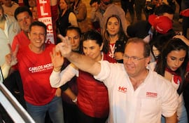 Camilo Pérez con gafas y camisa blanca, levantando la mano junto a Costanza Troche en chaqueta roja, sonrientes en un evento político.