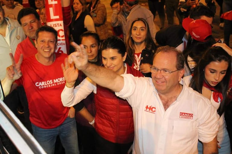 Camilo Pérez con gafas y camisa blanca, levantando la mano junto a Costanza Troche en chaqueta roja, sonrientes en un evento político.