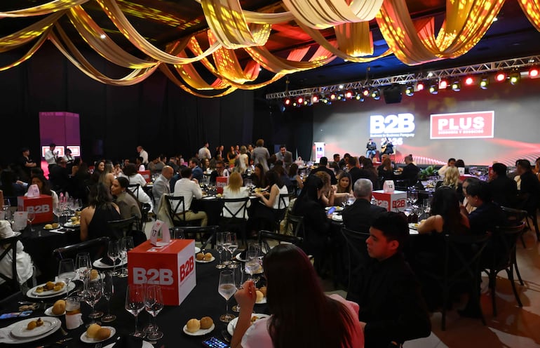 Mujer en el centro observa atentamente el escenario durante la cena de premiación, rodeada de mesas decoradas con copas y platos.