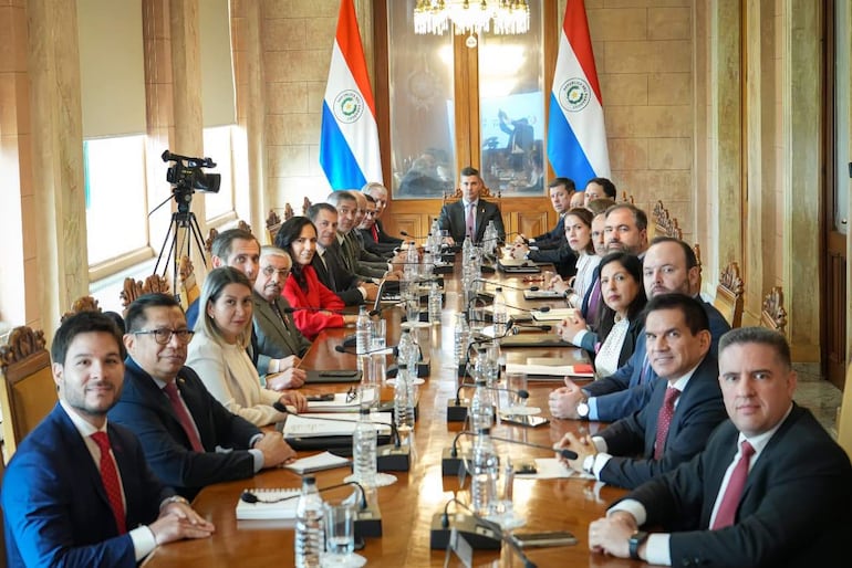 Santiago Peña en el centro, sentado erguido; asistentes formales escuchan atentamente en una sala con banderas de Paraguay.