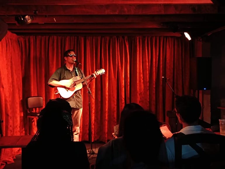Hombre joven toca guitarra acústica y canta en un escenario con iluminación tenue y fondo de cortinas rojas.