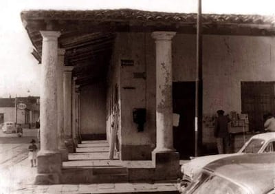 Automóvil antiguo estacionado frente a columnas clásicas. Persona al fondo junto a una mesa con mercancías.