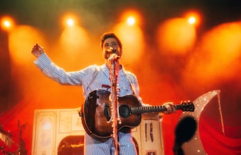 Músico en camisa de pijama de rayas, de pie con guitarra acústica bajo luces coloridas y ambiente vibrante.