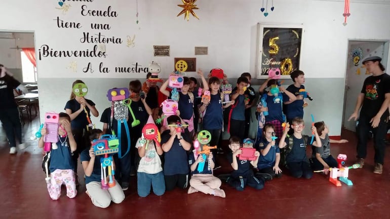 Veinticinco niños con camisetas oscuras sonríen mientras sostienen coloridos juguetes reciclados en un salón festivo.