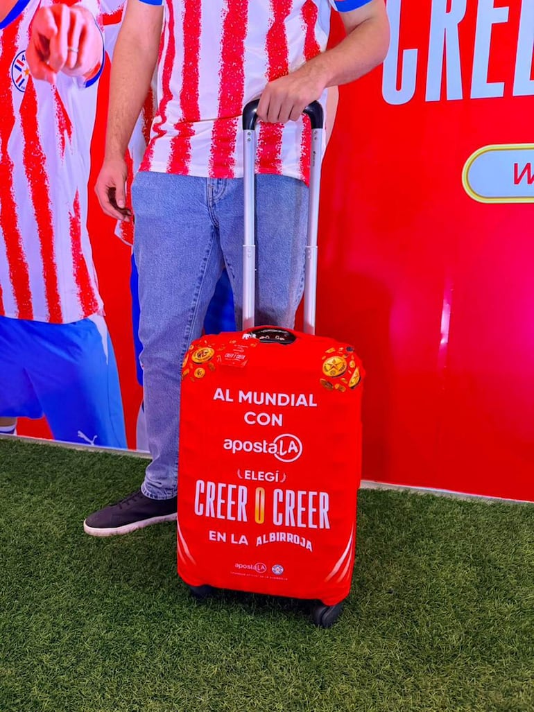 Hombre con camiseta a rayas rojas y blancas sostiene una maleta roja con inscripciones de apoyo a la Albirroja, frente a pared roja.