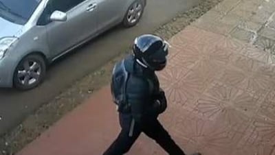 Persona con abrigo azul oscuro y casco, caminando cerca de un vehículo gris en un entorno urbano.