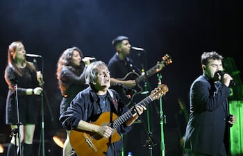 Dos cantantes con micrófonos y vestidos oscuros, un hombre cantando con guitarra en chaqueta negra, y un joven con traje oscuro en el escenario.