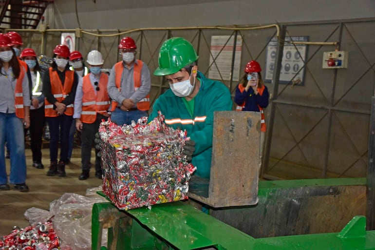 Hombre con casco verde y mascarilla opera una compactadora de latas, mientras otros observan con cascos y chalecos naranjas.
