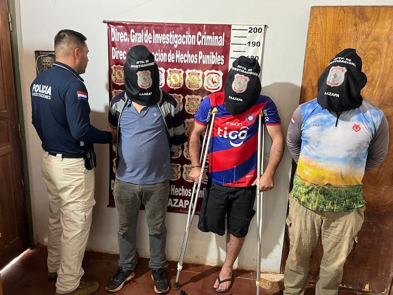 Tres hombres con capuchas oscuras, uno en muletas, conversan con un oficial de policía en una estación, con cartel visible de investigaciones criminales.