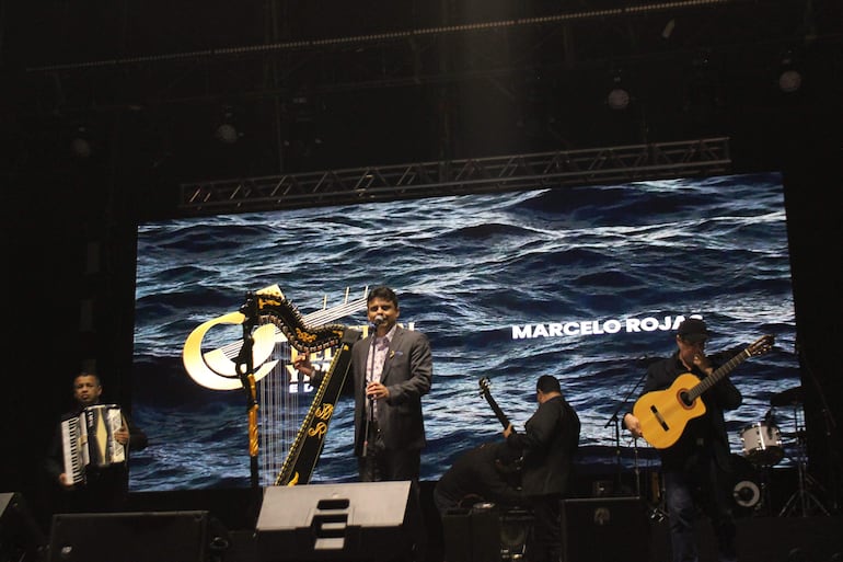 Cuatro músicos en el escenario: hombre con arpa en traje oscuro, acordeonista a su lado, guitarrista en segundo plano y músico de espaldas.