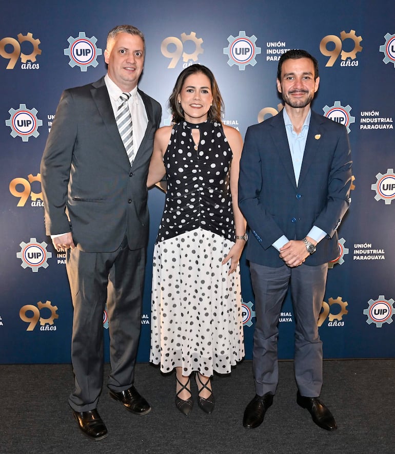 Hombre con traje gris habla, mujer sonriente con vestido a lunares centrales y hombre con traje oscuro sonriendo a la derecha.