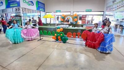 Cuatro niñas con trajes típicos coloridos bailan con alegría, en un ambiente festivo decorado con globos en tonos naranja y verde.