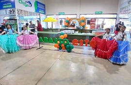 Cuatro niñas con trajes típicos coloridos bailan con alegría, en un ambiente festivo decorado con globos en tonos naranja y verde.