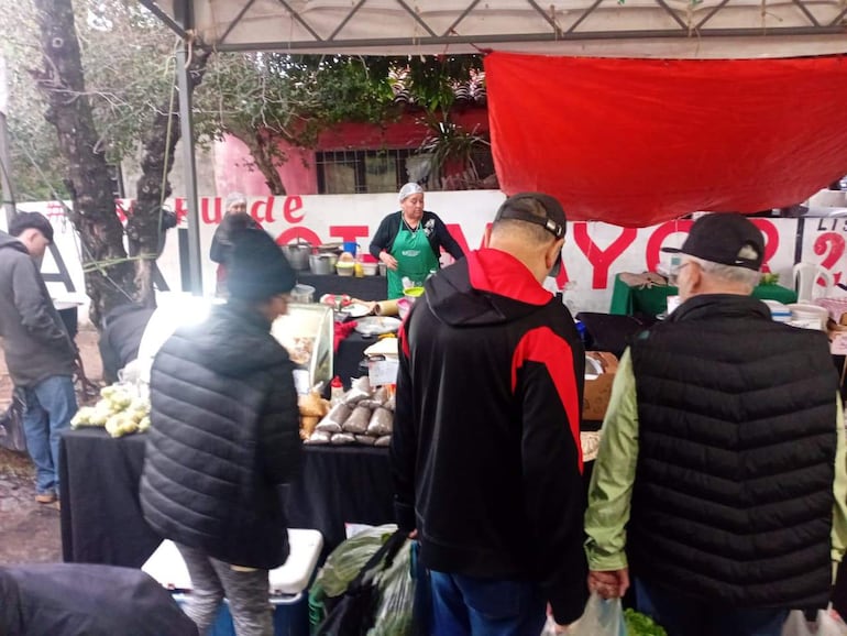 Mujer con delantal verde atiende un puesto en el mercado, mientras dos hombres eligen productos. Atmosfera activa y comunitaria.