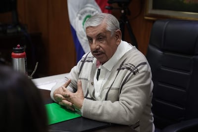 Isaías Ricardo Fretes, con gorra clara y suéter casual, habla mientras gesticula frente a una mesa con carpeta verde y termo metálico.