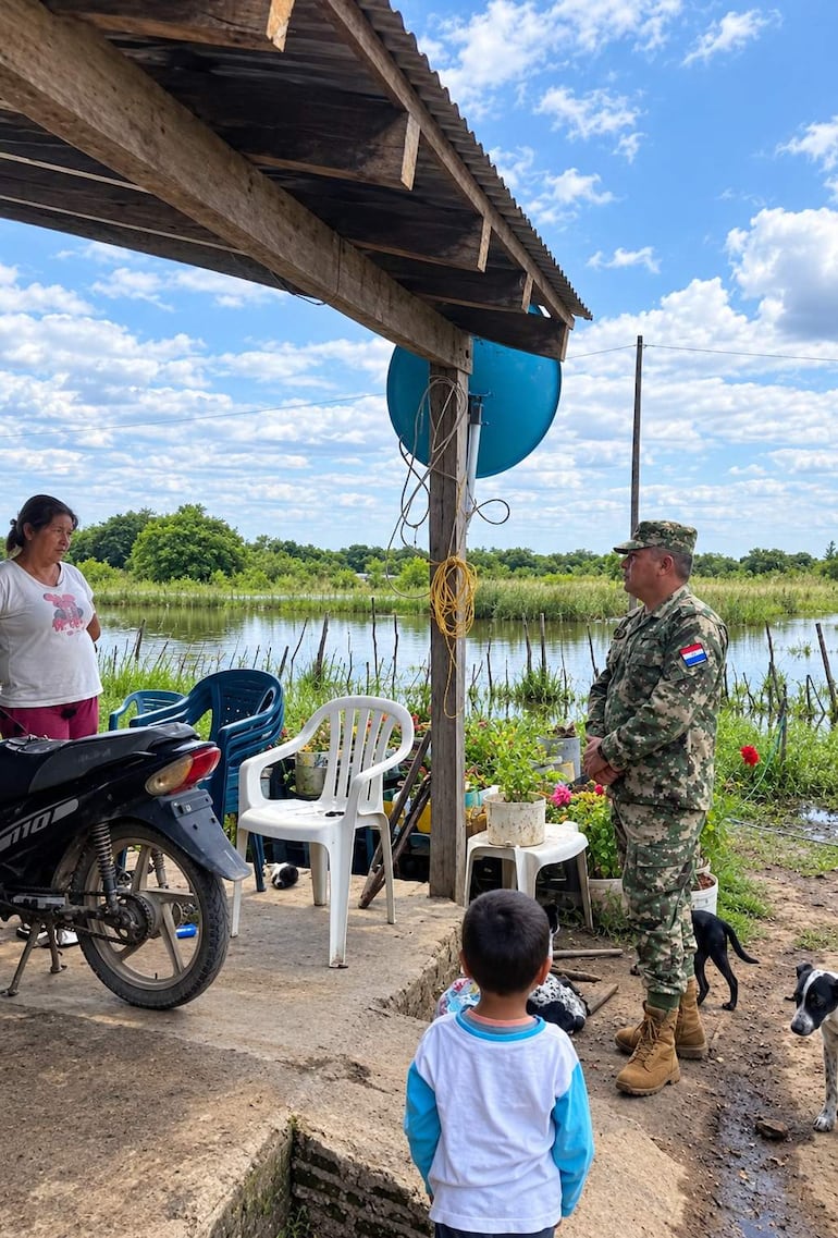 Mujer en camiseta blanca y pantalón rojo escucha a un soldado en uniforme camuflado, mientras un niño pequeño observa la conversación.