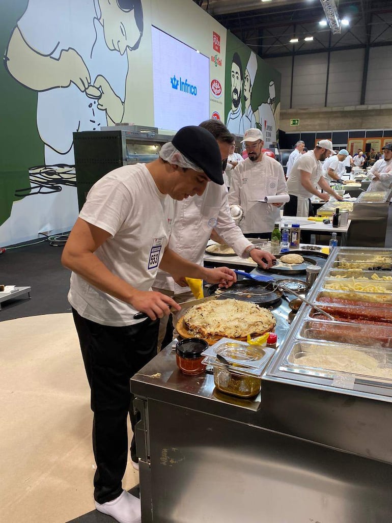 Chef en camiseta blanca y gorro negro decora pizza, mientras otros chefs trabajan en estaciones con ingredientes y utensilios.